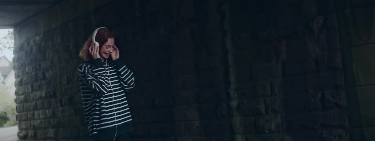 A young woman with red hair dancing and laughing while listening to music on her white headphones in an underpass