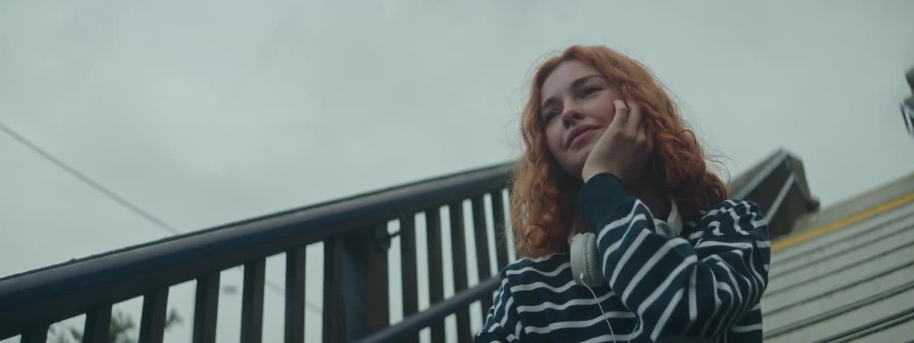 A young woman with flowing red hair sits on a staircase white headphones dangling around her neck