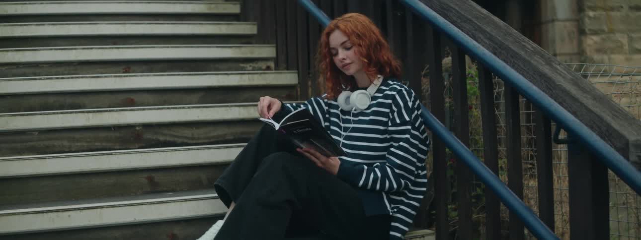 A red haired teenage girl reads a book while sitting on steps outside of a building