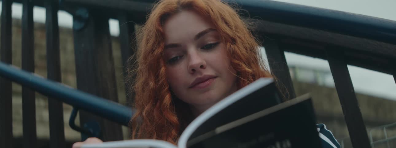A redheaded young woman sitting in a staircase reading a book