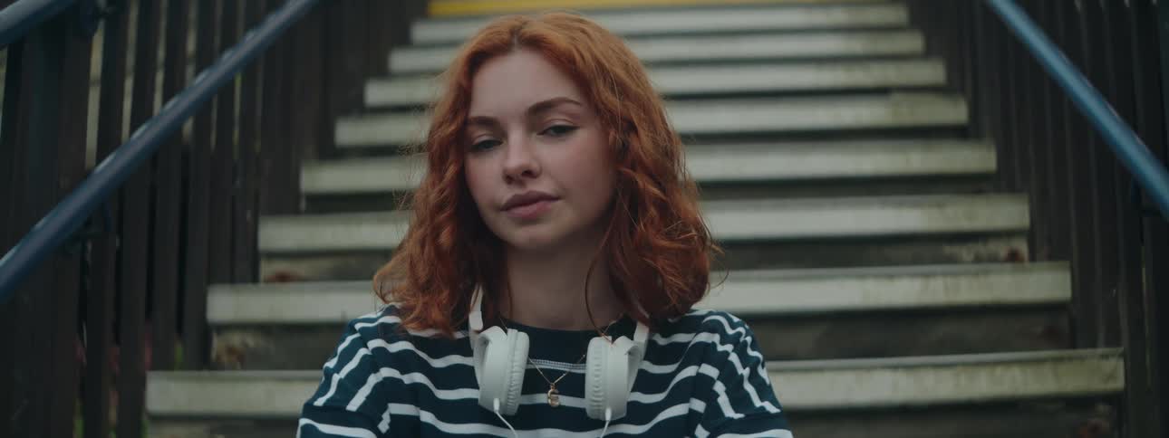 The face of a young woman with curly orange hair sitting on the steps of an outside staircase