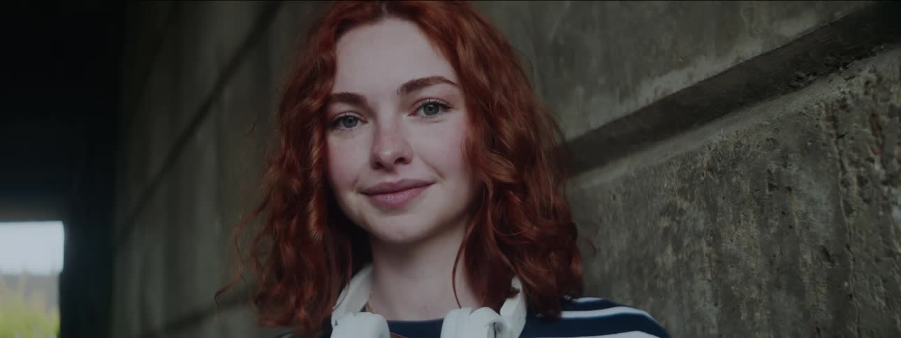 Redheaded woman leaning against a wall in a tunnel smiles