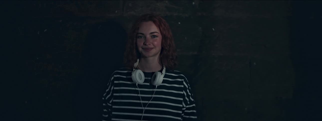 A young woman with headphones around her neck is looking straight ahead while giving a subtle smile
