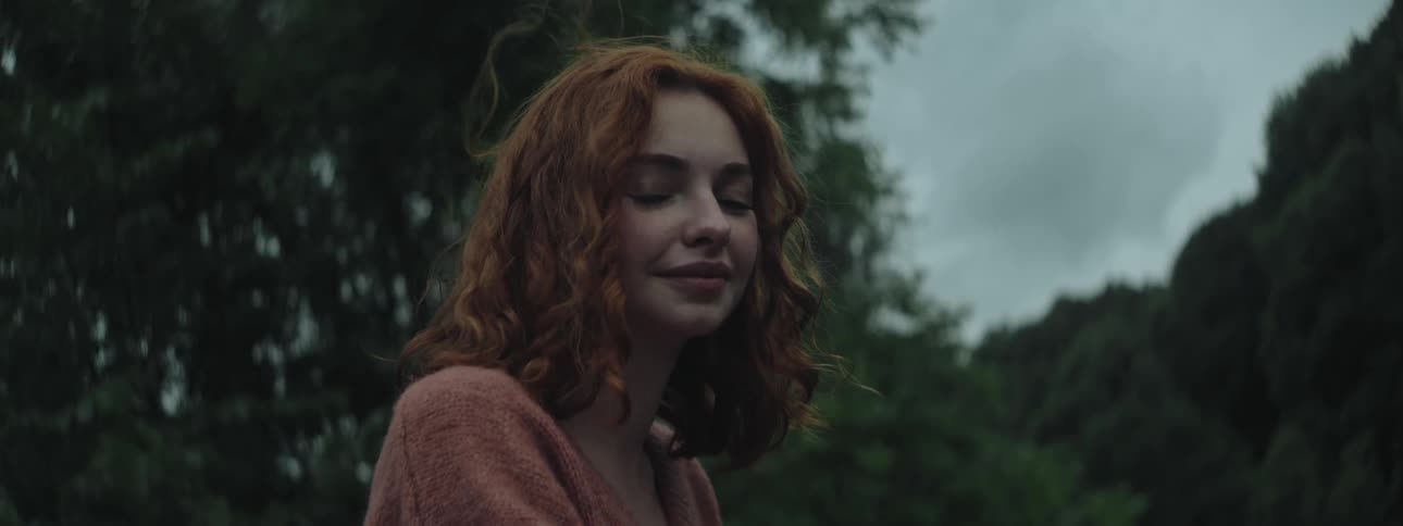 Smiling redheaded woman stands outdoors near the woods on a windy overcast day