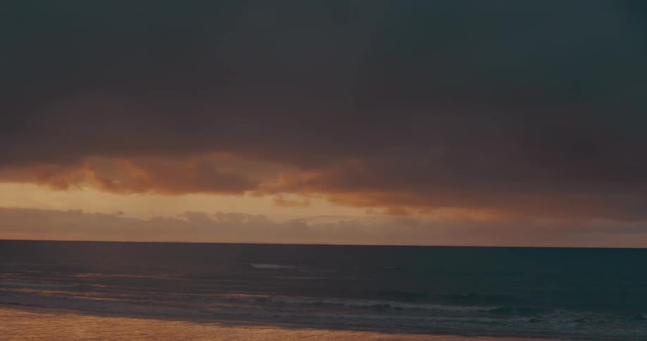 The setting sun casts a golden glow across the choppy waves as the sky above darkens with ominous clouds