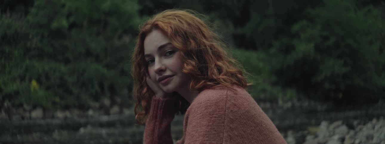 A redheaded young woman in a park by a river smiling while resting her face on her hand then turning her head