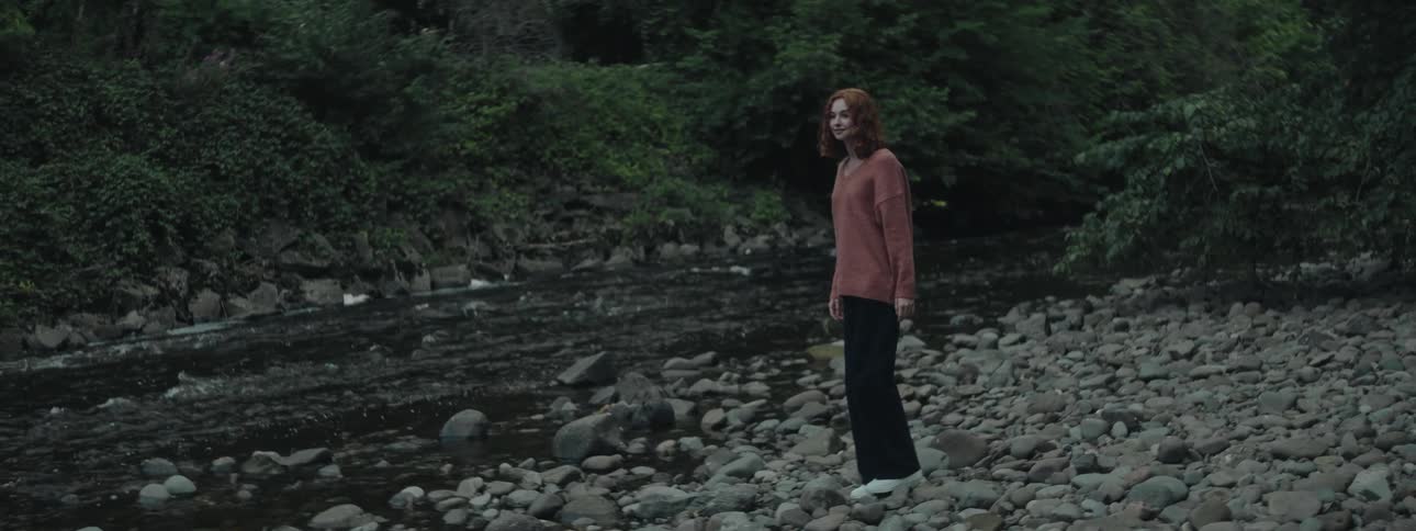 A young woman with red hair stands on the rocky shores of a river