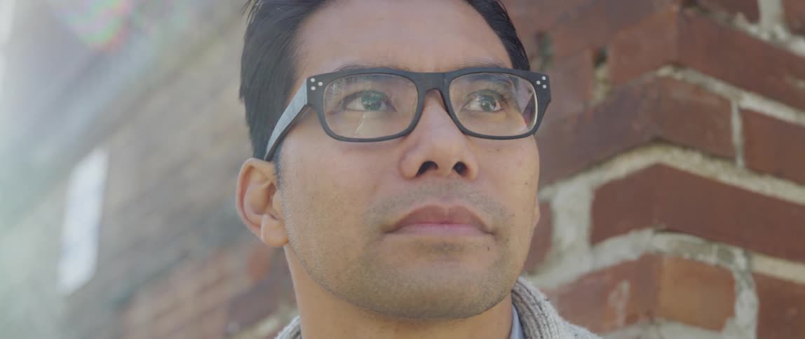 Man with glasses stands against a brick wall