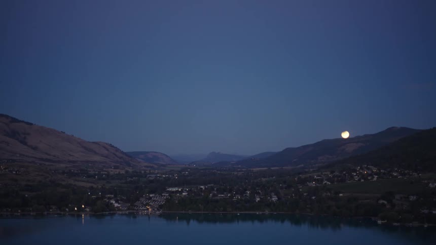 The moon rising over the mountains behind a city on the lake