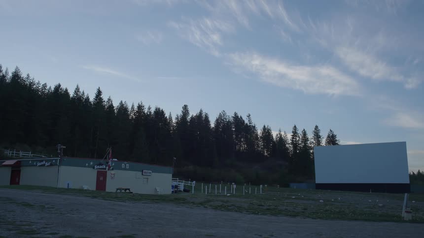 A field with some small white buildings and behind it there is a pine forest