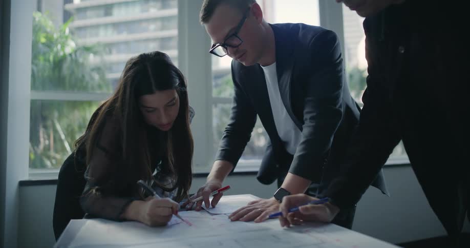 A group of three people are gathered around a table looking at documents and drawing on them