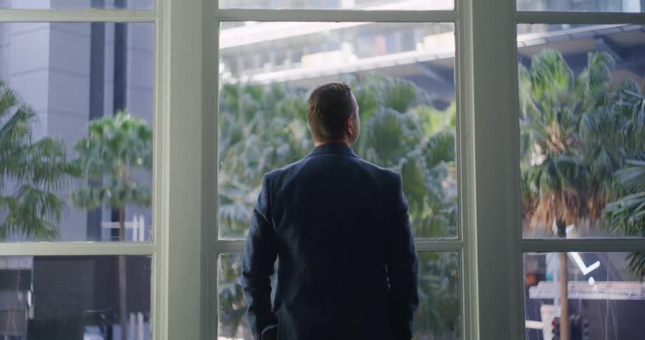 A man in a suit stands in front of a window overlooking a cityscape