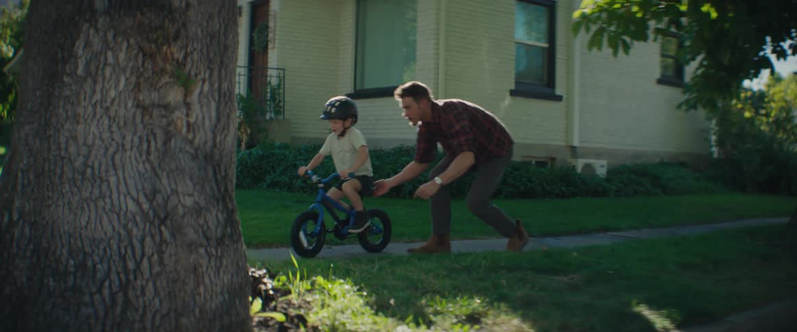 A father pushing his little son who is learning to ride a bike and he is wearing a helmet
