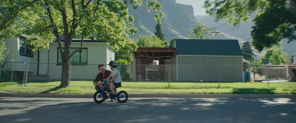 A father assists his son who is wearing a helmet while riding a bicycle on a residential street