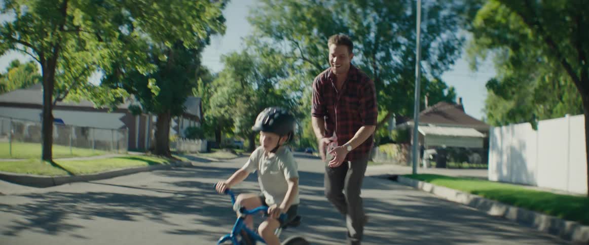 A father in a plaid shirt chases his young son who is riding a bicycle on the street