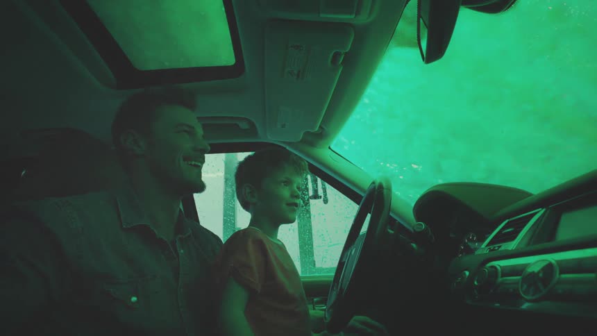The son is sitting on his fathers lap while they drive in the rain