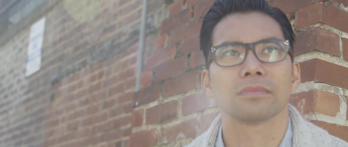 Man with glasses stands against a brick wall