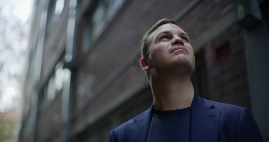A young man in a blazer stands outside a building and looks up