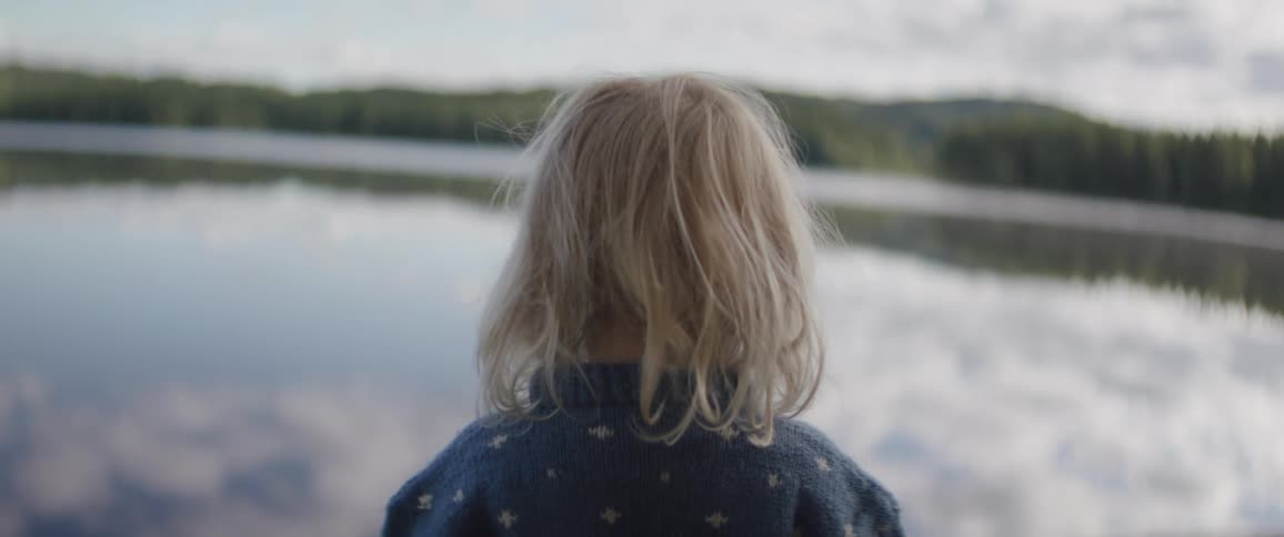 A young person in a blue sweater stands facing a body of water gazing at a duck in the foreground