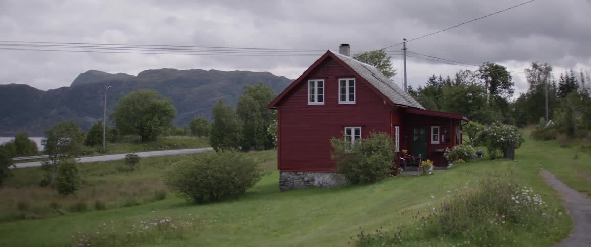 A small red house sits on a hill overlooking a valley with mountains in the distance