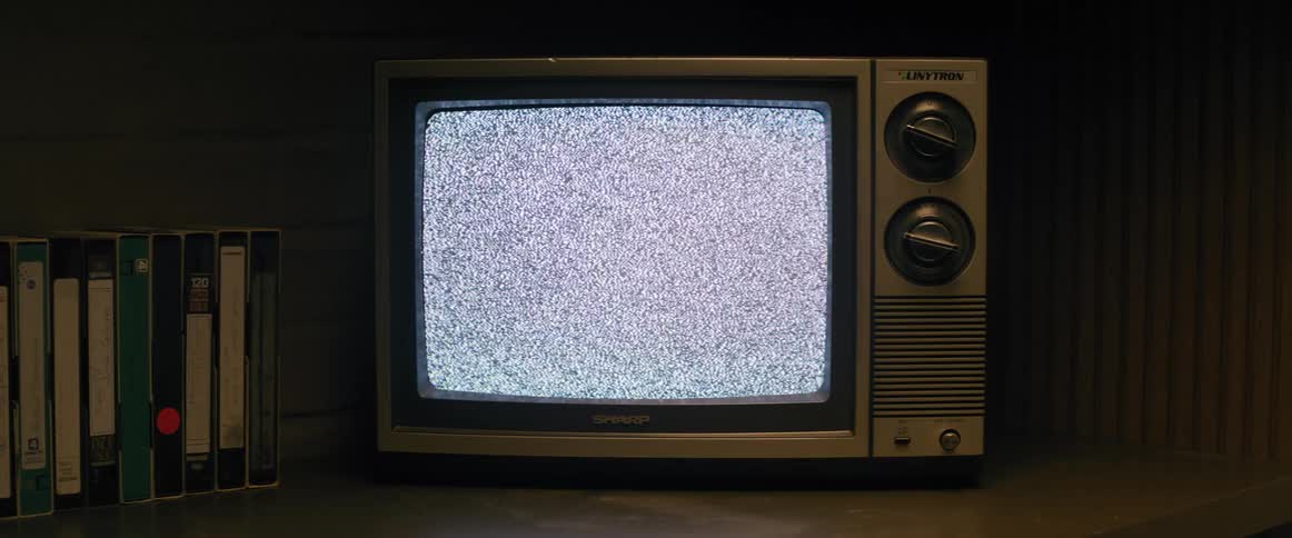 An old television with a lit screen with flashing black and white dots and a person walks in front of the television