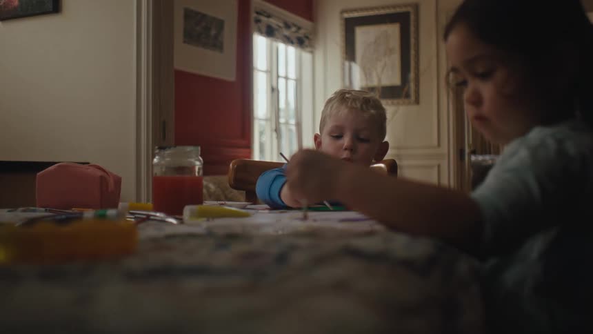 A little girl and a little boy are sitting around a table and are painting papers using brushes with paint