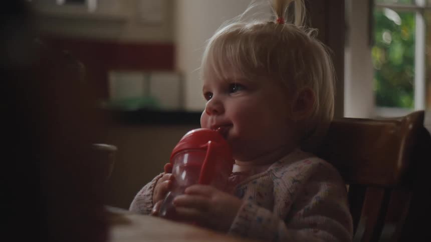 Little blonde girl sitting on a wooden chair in the kitchen drinking a bottle of water