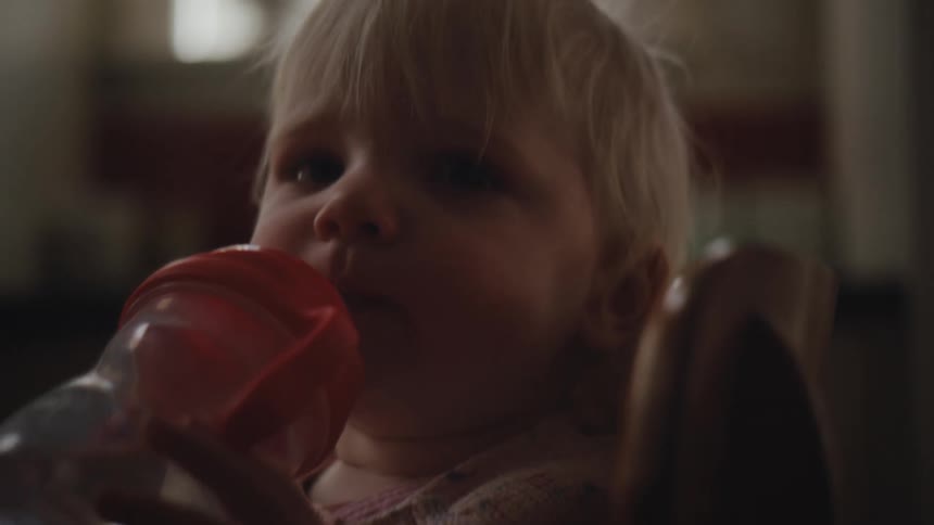 A young child with soft blonde hair drinking from a bottle in a dimly lit environment with the soft light highlighting their delicate features
