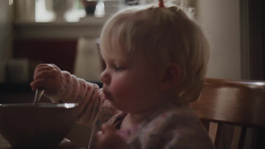 Little blonde girl sitting at the table holding a spoon and eating from a bowl