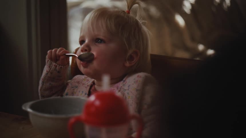 Little blonde girl sitting at the table eating with a spoon and a bowl