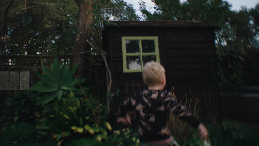 A little blond boy is running in a yard with green grass and there is a small wooden cabin behind