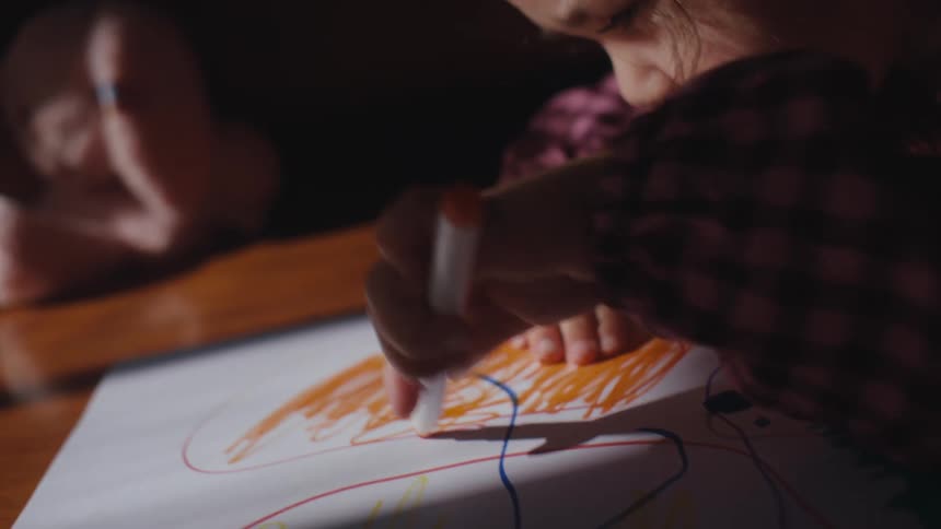 A young girl draws a picture with an orange marker while sitting at a wooden table