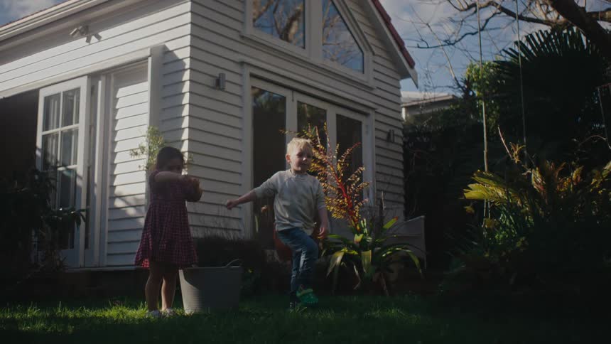 A little boy and a little girl are in a grassy yard and the little boy throws an object into the grass while jumping