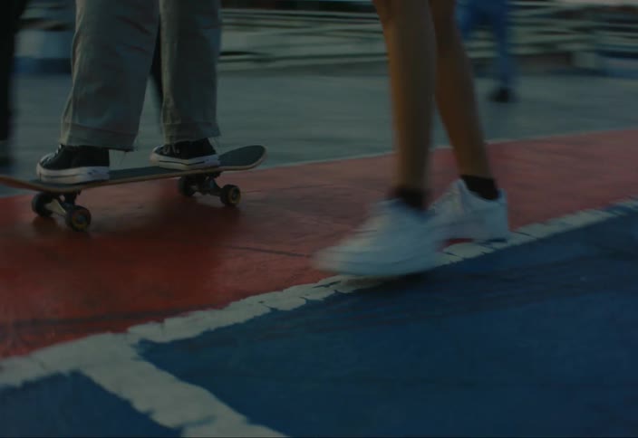 A closeup of a person riding a skateboard on a red and blue surface with other people walking nearby