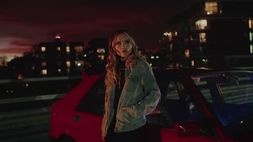 A woman standing beside a red car at night wearing a denim jacket with her hands in the pockets