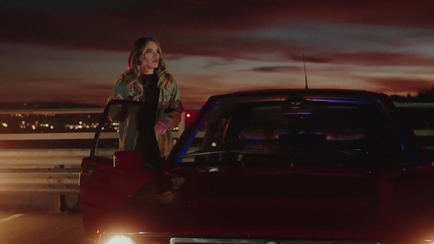 A blonde woman is getting into a red car that is parked at dusk
