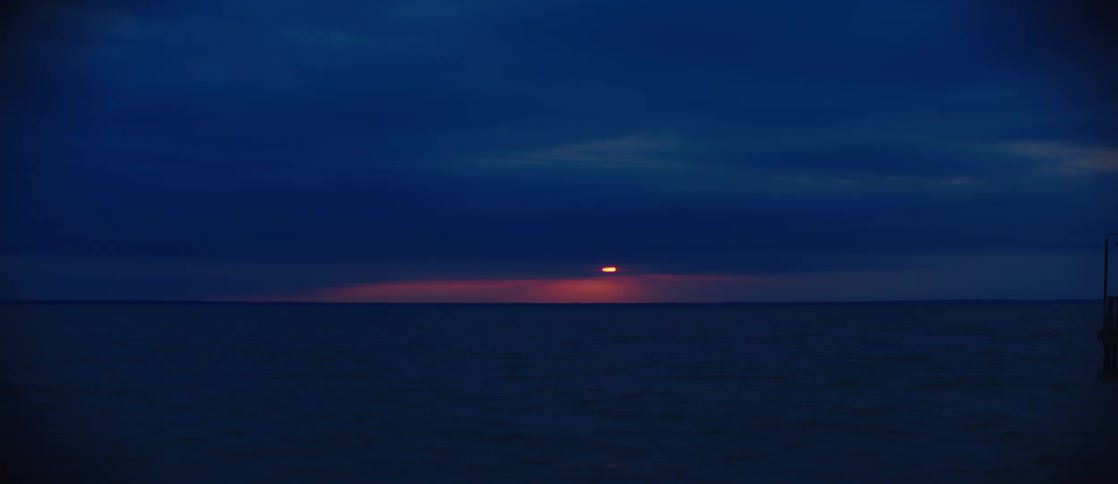 The sun sets over a calm vast ocean under a dark blue sky appearing as a small orange circle
