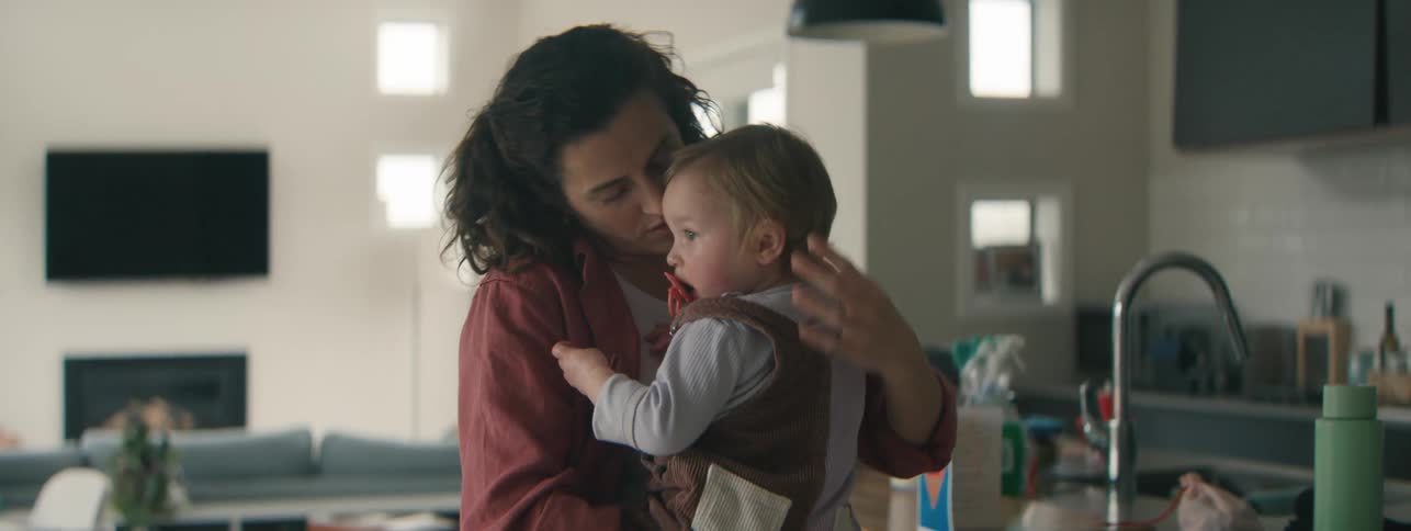 A mother holding a baby in her arms looking at the child affectionately while the baby gazes calmly ahead with a pacifier in its mouth set in a cozy softly lit home environment