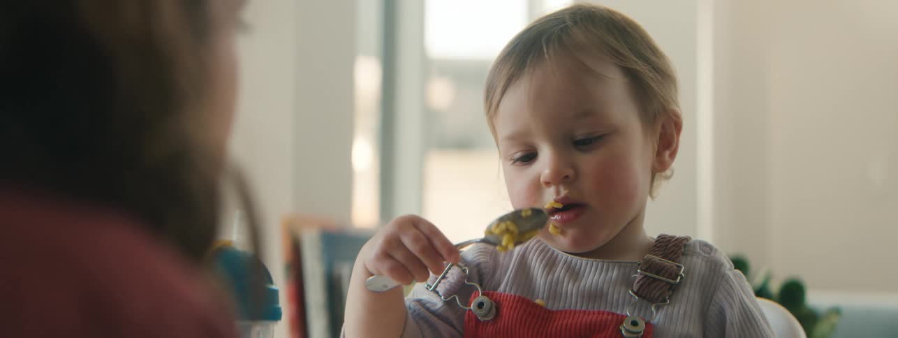 A young child wearing a striped shirt and red overalls is happily eating with a spoon gazing toward someone offscreen with a playful expression as they enjoy a meal in a bright softly lit room