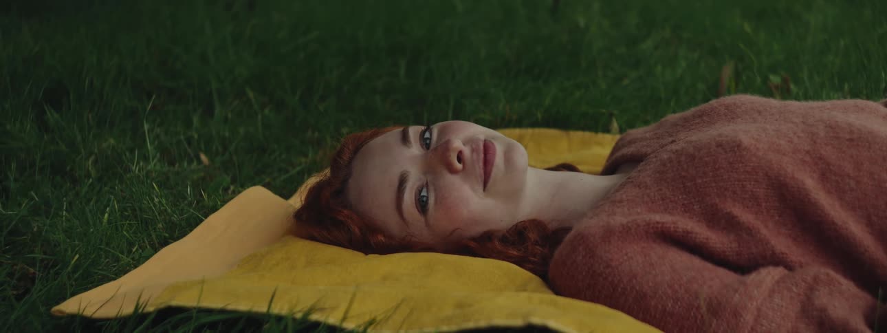 Redhead woman looking to the side with happy facial expression while lying on a cloth on the grass