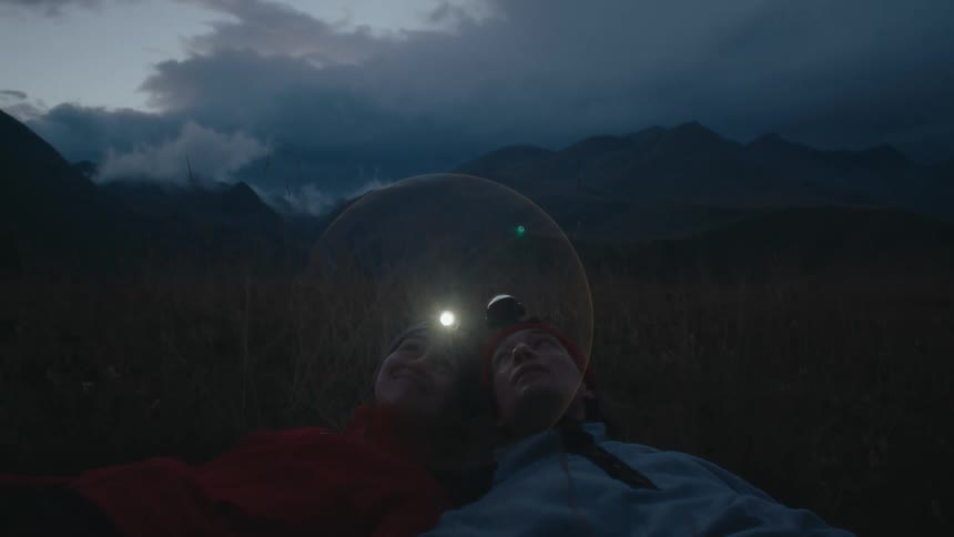 Man and woman with flashlight on their heads lying together on the grass with mountain ranges in the background on a cloudy day