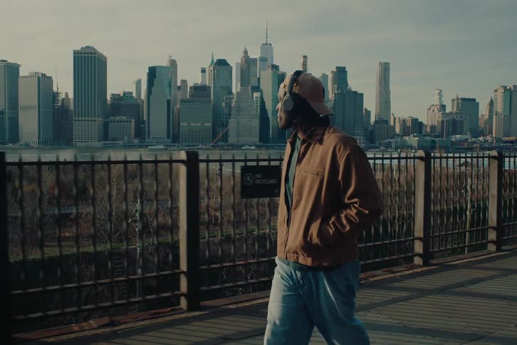 Man listening to music on headphones walks across a bridge across the water from the city skyline