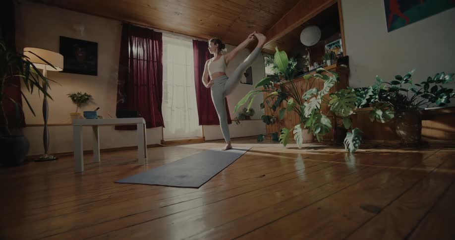 A young woman stands on a yoga mat in a living room to perform a leg stretch