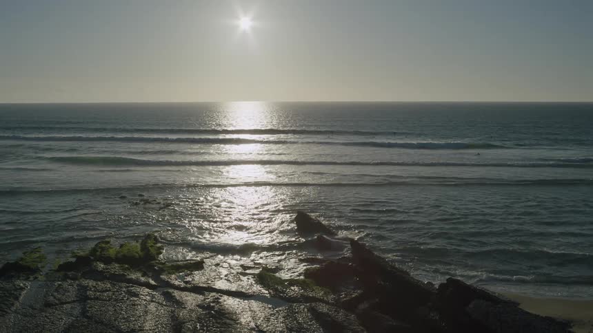 The sun hangs in the sky over an ocean and waves hitting a beach and rocky coastal area