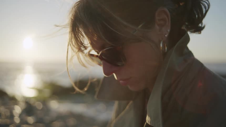 Portrait of a woman in a coat and sunglasses on the seashore with the sun on the horizon
