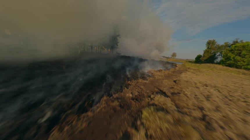 An FPV drone aerial view of a field on fire with smoke rising into the air
