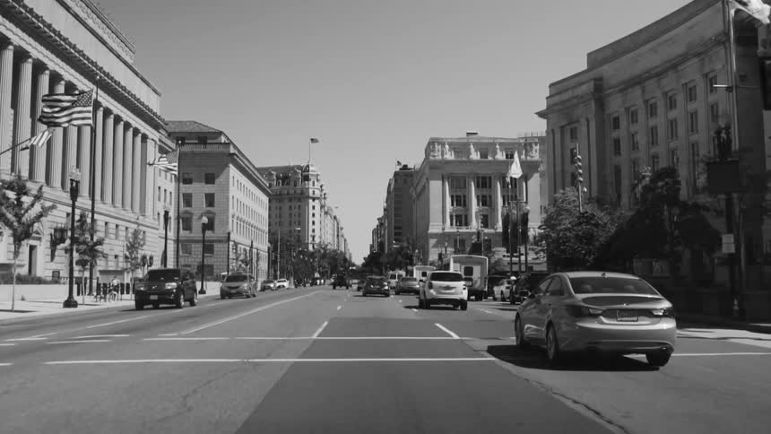 A black and white footage of a wide street with several cars driving down it