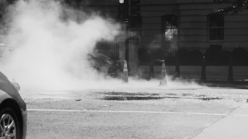 Traffic moves through a city next to steam emerging from the street