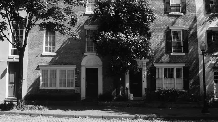 Black and white footage of a row of threestory brick townhouses with trees and a cobblestone street