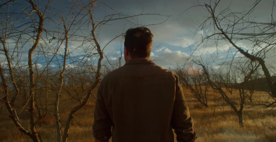 A man wearing a brown jacket stands on a hill scattered with driedup small trees
